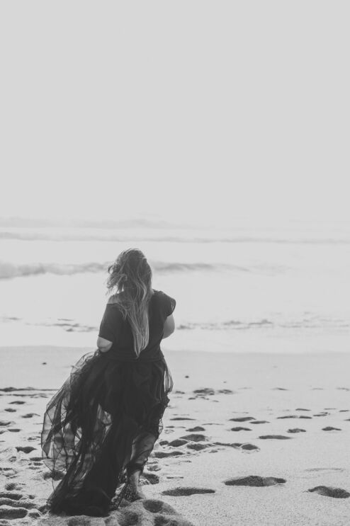 Uma mulher, de costas, na praia, usa um vestido comprido