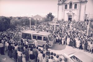 Multidão reúne-se para um funeral, com carro funerário e igreja visíveis