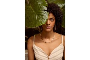 Mulher posa com colar e brincos dourados perto de uma grande folha verde