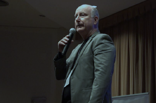 Homem discursa ao microfone num palco com cortinas