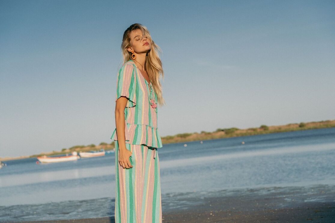 Mulher de cabelo loiro, de olhos fechados, usa um vestido às riscas coloridas numa praia