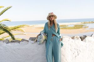 Modelo com chapéu de palha e conjunto de calças e camisa azul turquesa, junto ao mar